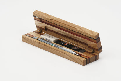 A wooden pencil case with an open hinged lid, showcasing a striped wood grain pattern in natural tones. Inside, a mechanical pencil with a silver barrel is nestled with replacement leads and eraser in custom compartments, all set against a clean white background.